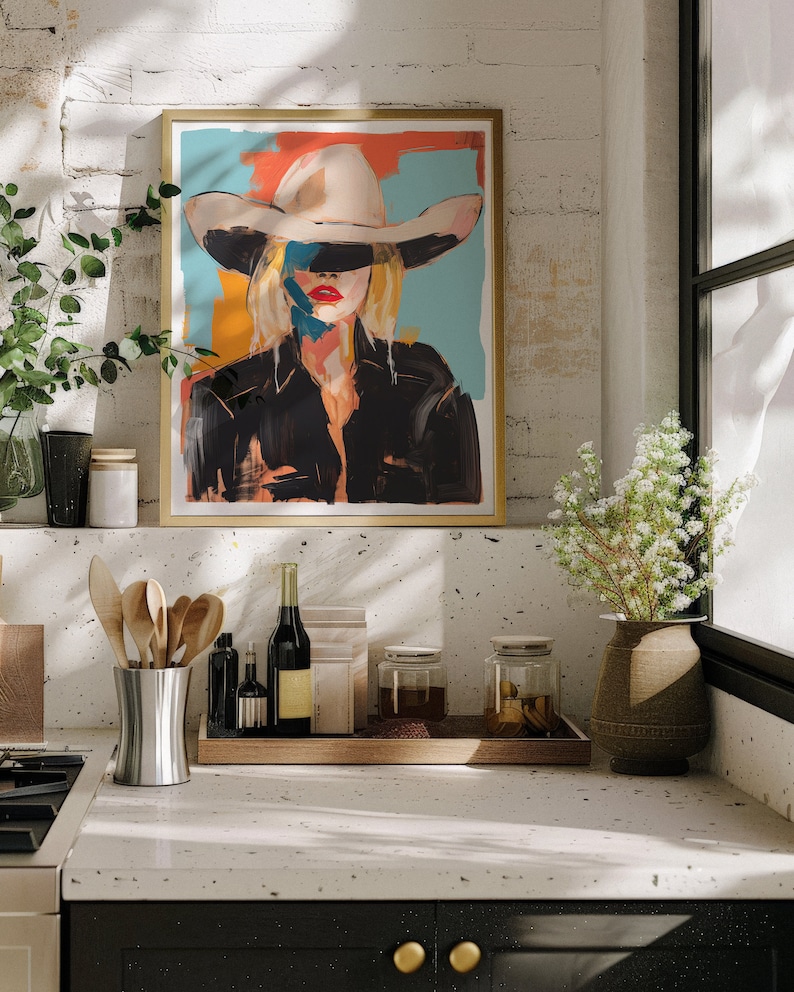 May include: A framed painting of a woman wearing a white cowboy hat and a black leather jacket. The background is a colorful abstract design. The painting is hanging on a white brick wall above a kitchen counter.
