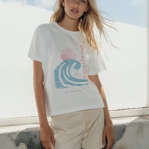 K&ouml;nnte beinhalten: Ein wei&szlig;es kurz&auml;rmeliges T-Shirt mit einem Grafikdruck einer blauen Welle, einer rosa Sonne und dem Text "POSITANO SURF" sowie "SURF CAMP ITALY 1980" am unteren Rand. Dazu kombiniert ist eine hellbeige Jeansshorts mit ausgefranstem Saum.