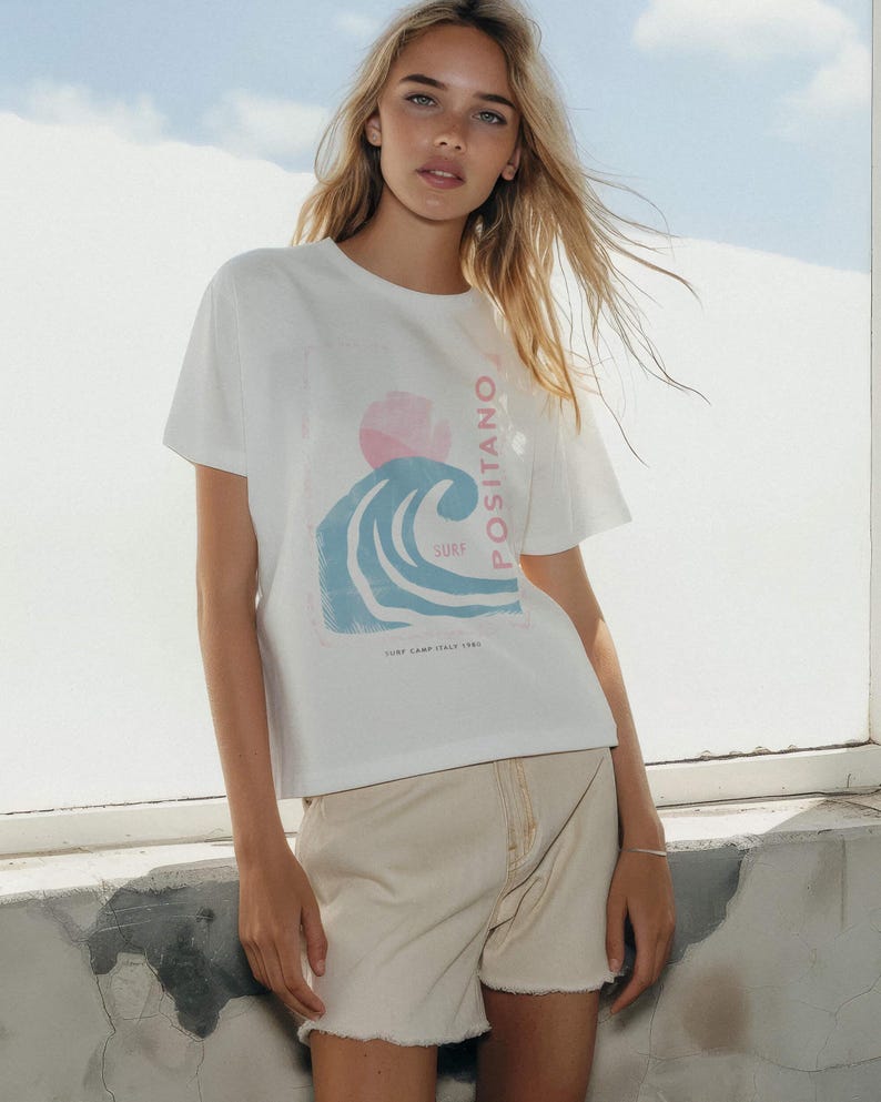 K&ouml;nnte beinhalten: Ein wei&szlig;es kurz&auml;rmeliges T-Shirt mit einem Grafikdruck einer blauen Welle, einer rosa Sonne und dem Text "POSITANO SURF" sowie "SURF CAMP ITALY 1980" am unteren Rand. Dazu kombiniert ist eine hellbeige Jeansshorts mit ausgefranstem Saum.