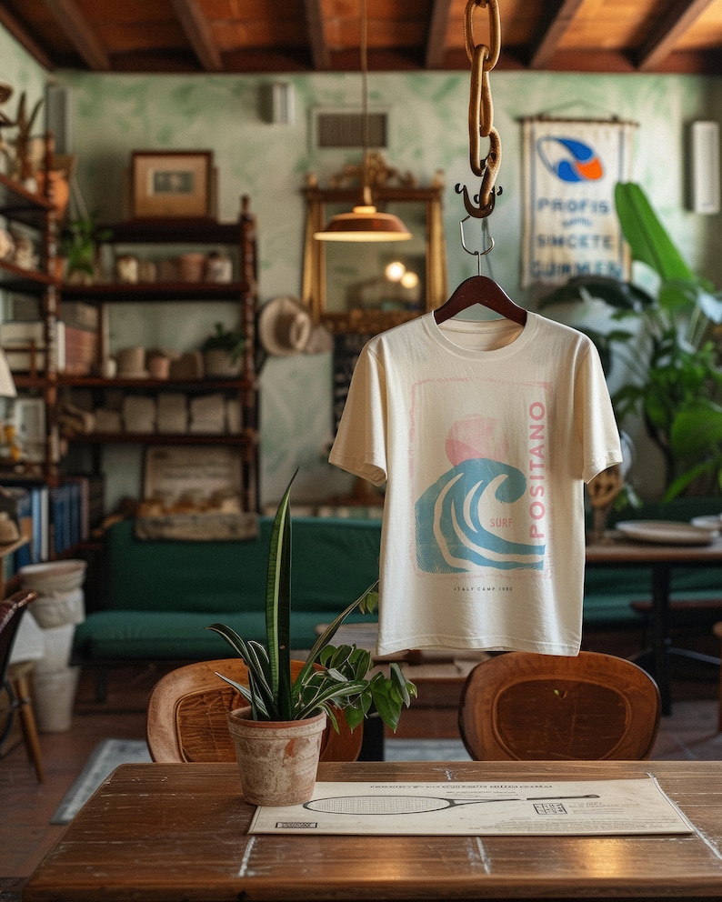 K&ouml;nnte beinhalten: Ein cremefarbenes Kurzarm-T-Shirt mit einem Grafikdruck einer blauen Meereswelle, einer rosa Sonne und dem vertikalen Text "POSITANO" und "SURF", wobei "ITALY CAMP 1980" unter dem Wellenmotiv auf der Vorderseite des Kleidungsst&uuml;cks gedruckt ist.