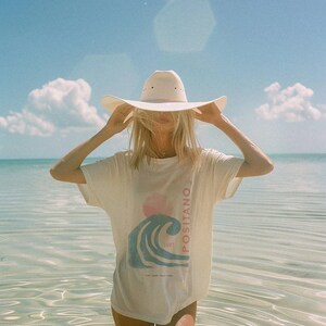 K&ouml;nnte beinhalten: Ein cremefarbenes Kurzarm-T-Shirt mit einem Grafikdruck einer blauen Welle, einer rosa Sonne und dem vertikalen Schriftzug "POSITANO" neben dem Wort "SURF" sowie dem kleineren Text "SURF CAMP ITALY 1989". Dazu wird ein breitkrempiger Hut getragen.