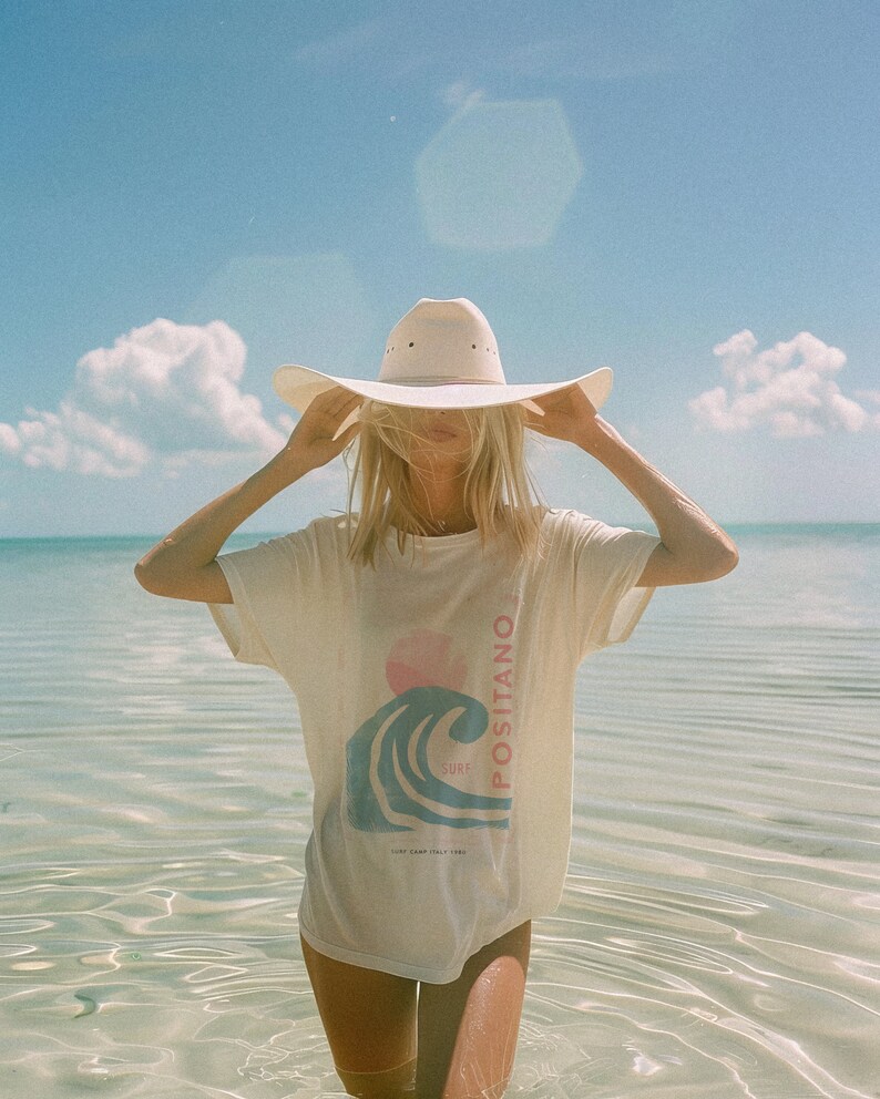 K&ouml;nnte beinhalten: Ein cremefarbenes Kurzarm-T-Shirt mit einem Grafikdruck einer blauen Welle, einer rosa Sonne und dem vertikalen Schriftzug "POSITANO" neben dem Wort "SURF" sowie dem kleineren Text "SURF CAMP ITALY 1989". Dazu wird ein breitkrempiger Hut getragen.