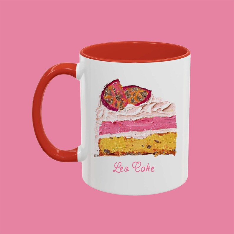 May include: White ceramic mug with a red rim and handle. The mug features a painting of a slice of cake with pink frosting and passion fruit on top. The text "Leo Cake" is printed below the cake.