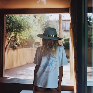 May include: A woman wearing a light blue hat with a gold band and a white t-shirt with a graphic of a cowboy riding a horse. The t-shirt says "Palm Springs Rodeo Club".