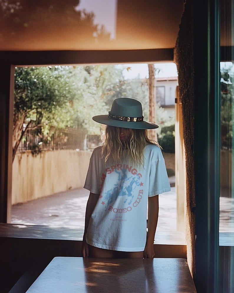 May include: A woman wearing a light blue hat with a gold band and a white t-shirt with a graphic of a cowboy riding a horse. The t-shirt says "Palm Springs Rodeo Club".