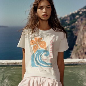 May include: A white short-sleeved t-shirt featuring a graphic print of an orange flower and a blue wave. The text on the shirt reads POSITANO SURF and ITALY CAMP 1980. The shirt is paired with a light pink pleated skirt with a flared hemline.