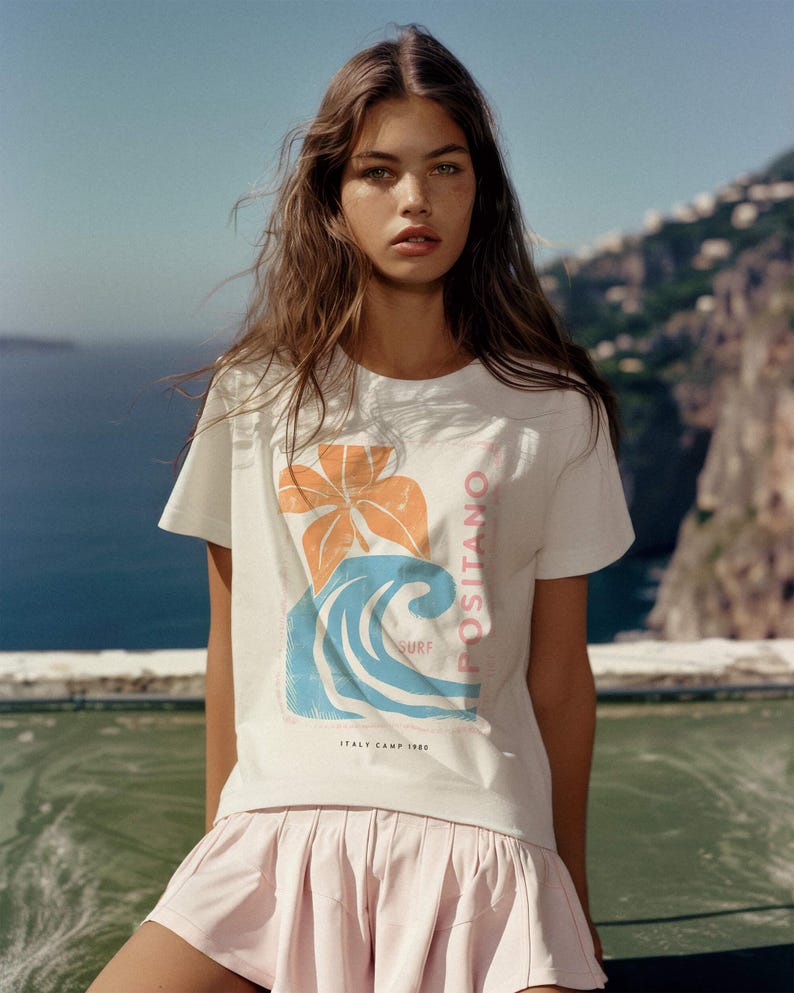 May include: A white short-sleeved t-shirt featuring a graphic print of an orange flower and a blue wave. The text on the shirt reads POSITANO SURF and ITALY CAMP 1980. The shirt is paired with a light pink pleated skirt with a flared hemline.