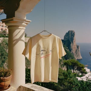 May include: A light yellow t-shirt with a graphic of a car driving on a winding road with the text "Amalfi Coast" in red. The t-shirt is hanging on a clothesline against a backdrop of a blue sky, a rocky cliff, and the ocean.