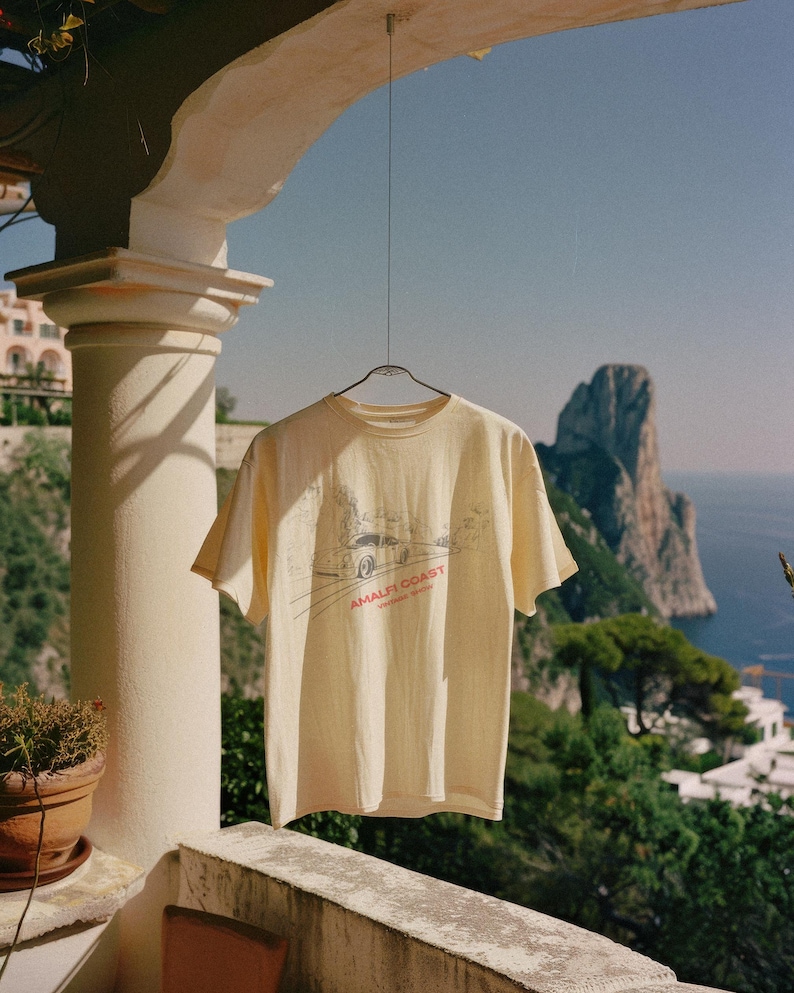 May include: A light yellow t-shirt with a graphic of a car driving on a winding road with the text "Amalfi Coast" in red. The t-shirt is hanging on a clothesline against a backdrop of a blue sky, a rocky cliff, and the ocean.