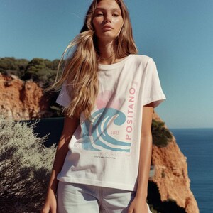 K&ouml;nnte beinhalten: Ein wei&szlig;es Kurzarm-T-Shirt mit einem Grafikdruck einer blauen Welle, einer rosa Sonne und dem vertikal an der Seite verlaufenden Schriftzug "POSITANO", sowie "SURF" und "SURF CAMP ITALY 1980" unter dem Wellenmotiv.