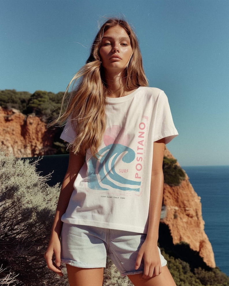K&ouml;nnte beinhalten: Ein wei&szlig;es Kurzarm-T-Shirt mit einem Grafikdruck einer blauen Welle, einer rosa Sonne und dem vertikal an der Seite verlaufenden Schriftzug "POSITANO", sowie "SURF" und "SURF CAMP ITALY 1980" unter dem Wellenmotiv.