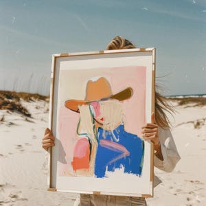 May include: A rectangular framed art print featuring an abstract painting of a figure wearing a tan cowboy hat. The artwork uses a colour palette of soft pink, bright blue, and orange brushstrokes against a light background, presented in a frame.
