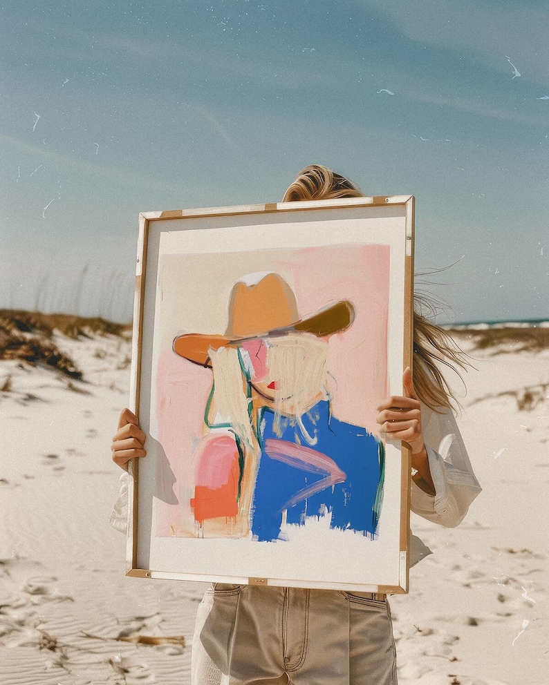 May include: A rectangular framed art print featuring an abstract painting of a figure wearing a tan cowboy hat. The artwork uses a colour palette of soft pink, bright blue, and orange brushstrokes against a light background, presented in a frame.