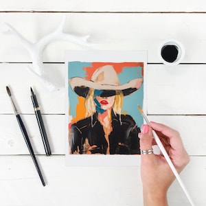 May include: A painting of a woman wearing a white cowboy hat with a blue, orange, and red background. The woman is wearing a black jacket and has blonde hair. The painting is being worked on with a paintbrush.