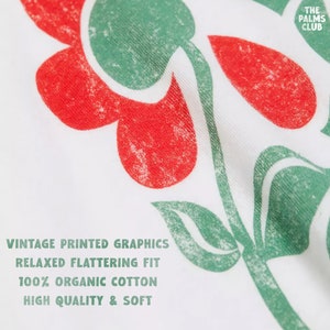 May include: White t-shirt with a red and green floral graphic print. The text "THE PALMS CLUB" is printed in the top right corner. The text "VINTAGE PRINTED GRAPHICS, RELAXED FLATTERING FIT, 100% ORGANIC COTTON, HIGH QUALITY & SOFT" is printed in the bottom left corner.