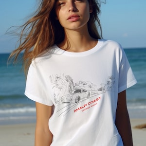 May include: White t-shirt with a black and white illustration of a car driving on a winding road with the text "Amalfi Coast Vintage Show" in red and black.