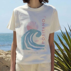 K&ouml;nnte beinhalten: Ein cremefarbenes Kurzarm-T-Shirt mit einem Grafikdruck einer stilisierten blauen Meereswelle, einer rosa Sonne und dem Text POSITANO, SURF und SURF CAMP ITALY 1980. Das Design ist mittig auf der Brust des Rundhals-T-Shirts platziert.