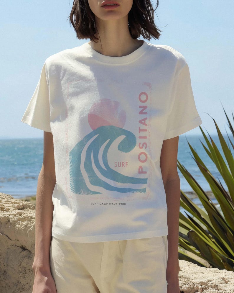 K&ouml;nnte beinhalten: Ein cremefarbenes Kurzarm-T-Shirt mit einem Grafikdruck einer stilisierten blauen Meereswelle, einer rosa Sonne und dem Text POSITANO, SURF und SURF CAMP ITALY 1980. Das Design ist mittig auf der Brust des Rundhals-T-Shirts platziert.