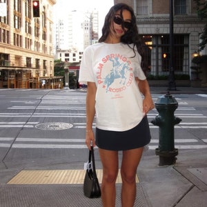 May include: A woman wearing a white t-shirt with a blue and red graphic that says "Palm Springs Rodeo Club 1980 California" and a black mini skirt. She is wearing white sneakers and a black handbag.