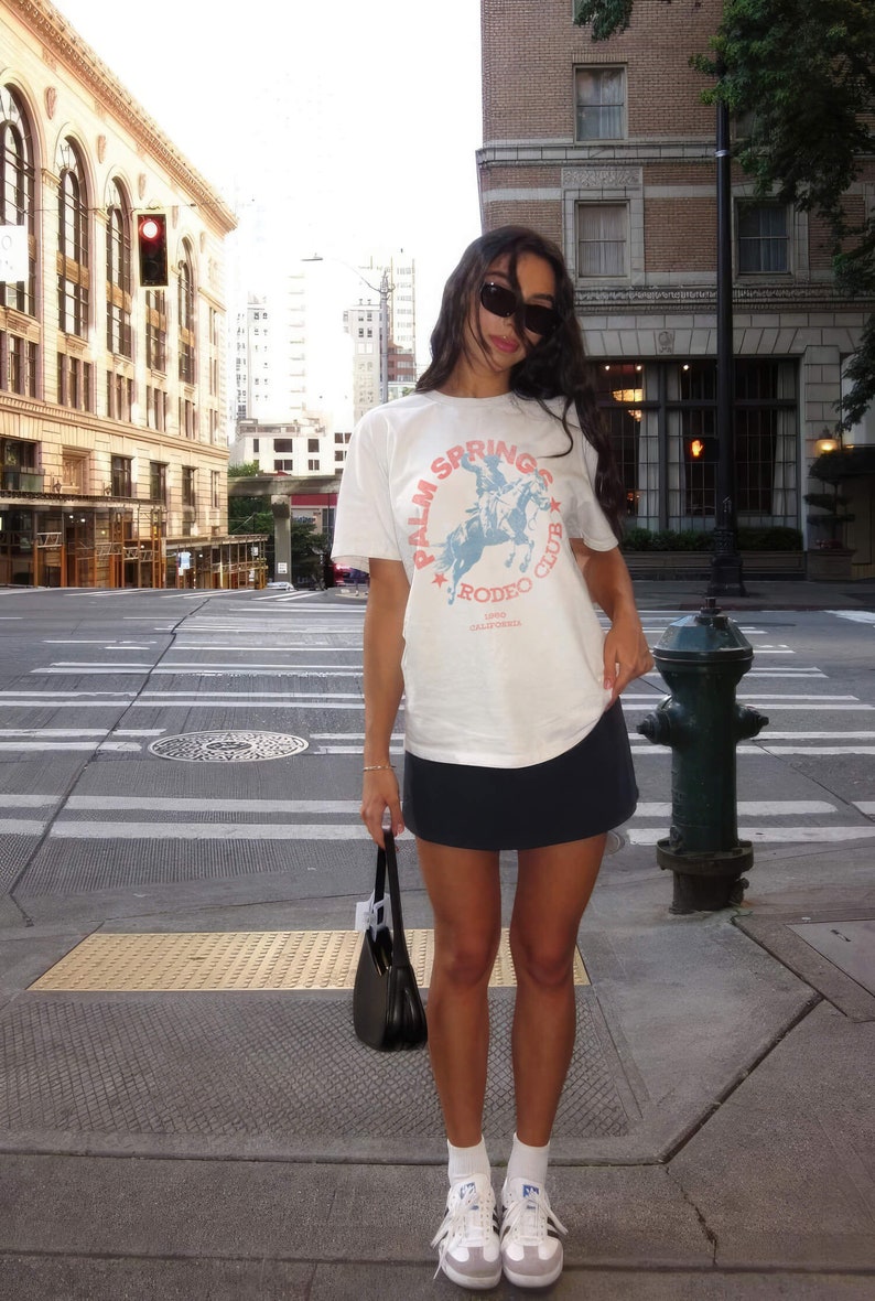 May include: A woman wearing a white t-shirt with a blue and red graphic that says "Palm Springs Rodeo Club 1980 California" and a black mini skirt. She is wearing white sneakers and a black handbag.