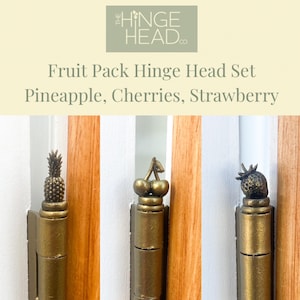May include: A set of three gold-toned hinge heads featuring a pineapple, cherries, and a strawberry. The hinge heads are mounted on wooden doors.