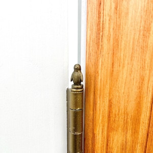 May include: A decorative brass door hinge featuring a small penguin figurine on top. The hinge is mounted on a white door frame and a wooden door with a natural wood grain finish.