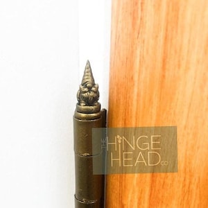 May include: A gold-toned metal hinge with a decorative gnome-shaped finial. The hinge is attached to a wooden door.