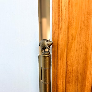 May include: A brass-colored metal door hinge topper featuring a small, sculpted figure of a pig standing on a circular base. The decorative ornament is attached to the top of a cylindrical hinge pin, finished with a metallic, aged patina texture.