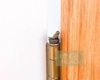 May include: A gold-toned door hinge with a small bird figurine perched on top. The bird is sitting in a nest and is facing to the right. The hinge is attached to a wooden door frame.