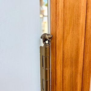 May include: A metallic gold-toned door hinge featuring a small, sculpted animal figure resembling a pig or boar perched on the top finial. The hinge is mounted vertically between a white wall and a vertical wooden door frame with visible grain.