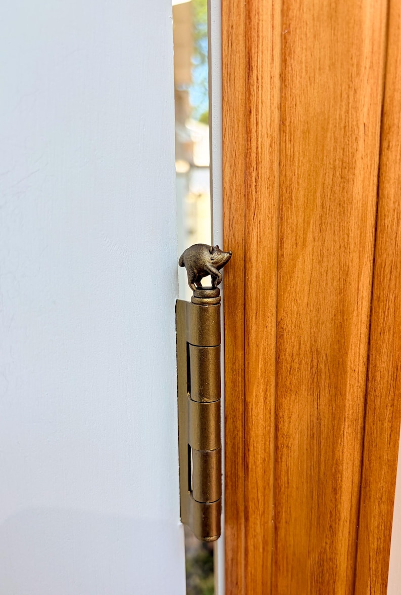 May include: A metallic gold-toned door hinge featuring a small, sculpted animal figure resembling a pig or boar perched on the top finial. The hinge is mounted vertically between a white wall and a vertical wooden door frame with visible grain.