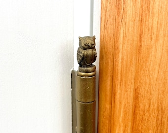 May include: A gold-toned door hinge with an owl finial. The owl is perched on a round base and has detailed feathers.