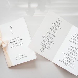 May include: A wedding ceremony program booklet with a simple floral design on the cover. The program lists the order of events for the wedding ceremony, including the entrance, liturgy of the word, and liturgy of the Eucharist. The wedding is for Elise Longhorn and David Jackson, and is taking place on October 11, 2023 at the Lourdes Catholic Church.