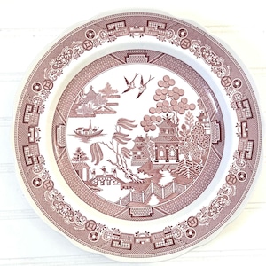 May include: A round, white and maroon plate with a scalloped edge. The plate features a detailed Willow pattern scene with pagodas, trees, bridges, and birds. The border has a repeating geometric and floral design.