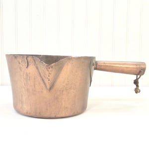 May include: A copper-colored metal pitcher with a wooden handle. The pitcher has a spout and a small rope tied to the end of the handle. The metal has a worn, aged appearance. The pitcher is likely used for pouring liquids.