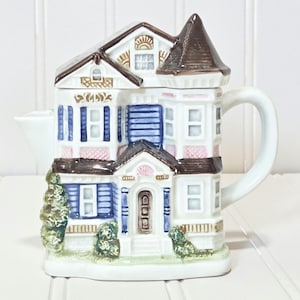 May include: A white ceramic teapot shaped like a Victorian house. The house-shaped teapot features blue shutters, pink accents, and a brown roof. The teapot has a white handle and spout, and is decorated with small green bushes.