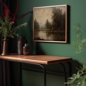Puede incluir: Una mesa consola de madera con un marco de metal negro, que presenta una pintura enmarcada de un paisaje de un lago y &aacute;rboles, un jarr&oacute;n marr&oacute;n y un frasco de vidrio en la superficie. La mesa est&aacute; colocada contra una pared verde oscuro.