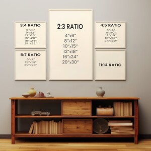 May include: A white wall with six framed posters showing different aspect ratios and their corresponding dimensions in inches. The posters are arranged in two rows of three. The bottom row has a poster with the text "11:14 RATIO". Below the posters is a wooden shelf unit with two shelves. The top shelf has a small brown vase and a small white ceramic object. The bottom shelf has a collection of books and a clock.