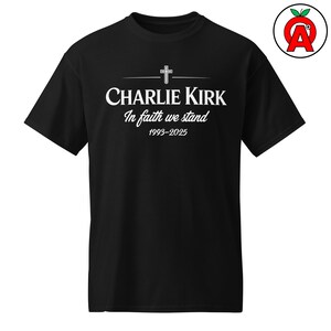 May include: Black t-shirt with white text. The text includes "CHARLIE KIRK" above "In faith we stand" and the years "1993-2025". A cross graphic is above the name. The shirt has short sleeves and a crew neck.