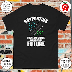 May include: Black t-shirt with the text "SUPPORTING LOCAL MACHINES THAT PREDICTS THE FUTURE" in white. The design includes blue and green dots with lines. Additional text includes "ASK ABOUT CUSTOM DESIGNS!" and "MORE COLOR OPTIONS!"