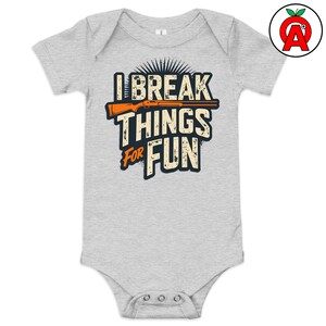 Könnte beinhalten: Ein hellgrauer Baby-Body mit dem Text "I BREAK THINGS FOR FUN" in einer Used-Schriftart. Eine orangefarbene Gewehrgrafik befindet sich über den Wörtern "THINGS" und "FOR". Der Body hat kurze Ärmel und einen Druckknopfverschluss.