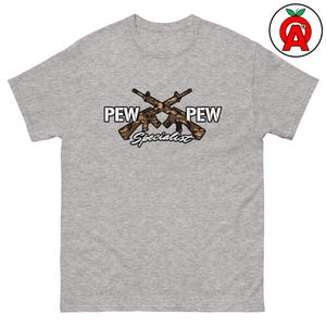Può includere: T-shirt grigia con la scritta "PEW PEW Specialist" e fucili incrociati con un motivo mimetico. La grafica è stampata sulla parte anteriore della maglietta.