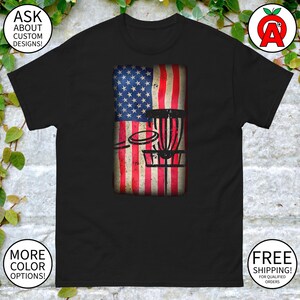 以下が含まれることがあります： アメリカ国旗のデザインが施された黒のTシャツ。その前にディスクゴルフのバスケットがある。