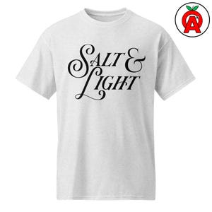 May include: A light gray t-shirt with the words "Salt & Light" in elegant black script. The shirt has short sleeves and a crew neck. A red apple logo with the letter "A" is in the upper right corner.