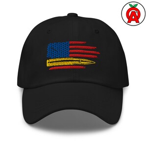 Op de afbeelding: Zwarte baseballcap met een geborduurd ontwerp van de Amerikaanse vlag en een gele kogel. Het vlagontwerp bevat blauwe sterren, rode strepen en een gele kogel. De cap is een klassieke, verstelbare stijl.