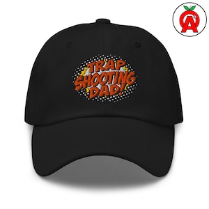 Könnte beinhalten: Schwarze Baseballkappe mit dem gestickten Text "TRAP SHOOTING DAD!" in Orange und Weiß mit gelben Akzenten. Der Text ist in einer Comic-Schriftart gehalten. Die Kappe hat einen gebogenen Schirm und einen Knopf oben.