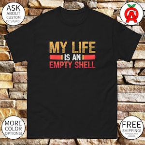 May include: Black t-shirt with the text "My Life Is An Empty Shell" in white and orange letters. The text is surrounded by two red and orange shotgun shells.