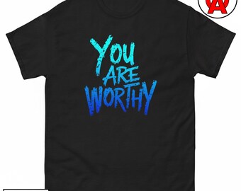 May include: A black short-sleeved t-shirt featuring the text "YOU ARE WORTHY" in a distressed, brush-stroke font. The lettering transitions from a light cyan at the top to a deep royal blue at the bottom. The shirt has a crew neckline.