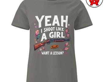 Puede incluir: Camiseta gris con el texto "YEAH, I SHOOT LIKE A GIRL, WANT A LESSON?" y un gráfico de una escopeta y un plato de arcilla. El diseño incluye detalles florales y de plumas.