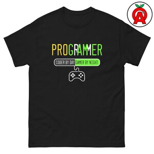 May include: A black short-sleeve t-shirt featuring the text PROGRAMMER in yellow and green. Below, a rectangular graphic reads CODER BY DAY GAMER BY NIGHT with a white line drawing of a game controller connected by a cord to the text box.